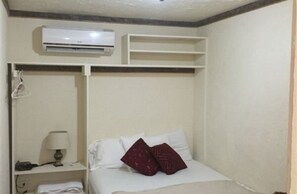 A small, single-bed room with a white bed, a red pillow, a bedside lamp, a white air conditioning unit, and a shelf above the bed. - Driftwood Gardens (Savaneta)