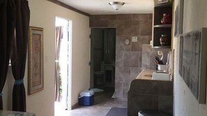Una cocina compacta con horno integrado, un lavamanos y una pequeña encimera. Se ve una puerta que da a un baño, y un cubo de basura en el suelo.