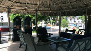 Un patio exterior con mobiliario de mimbre, una parrilla y vistas a una piscina y edificios al fondo.