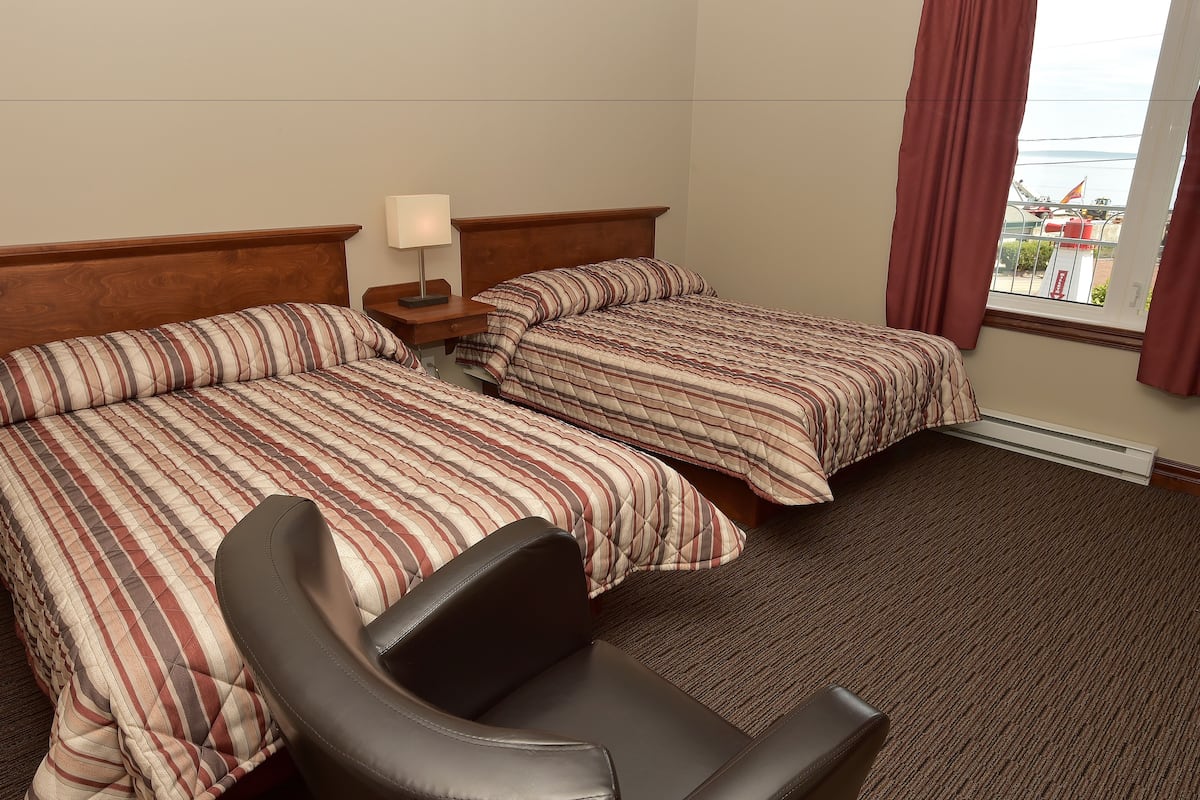 A hotel room with two beds, a chair, a nightstand with a lamp, and a window with curtains.