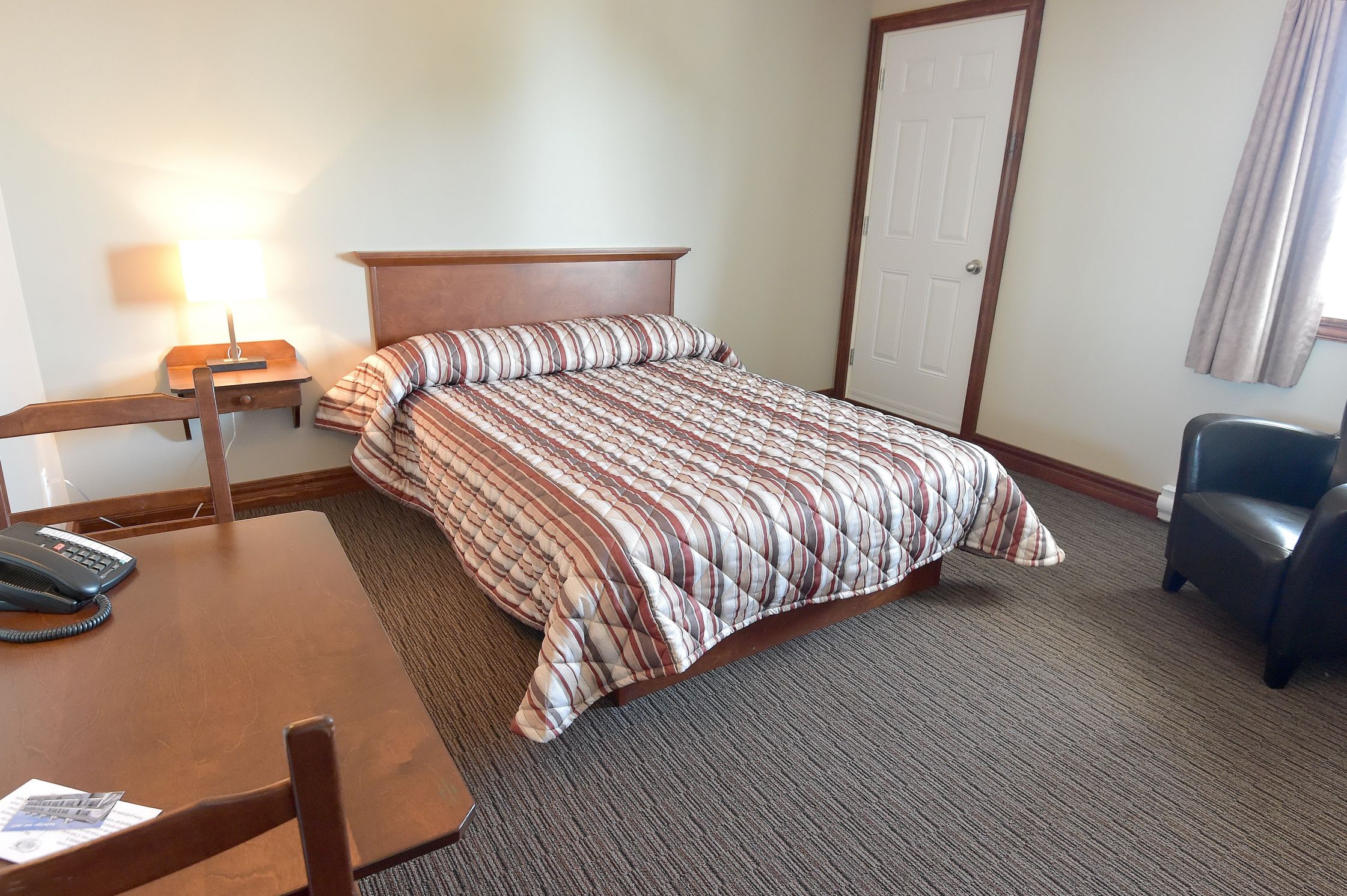 Une chambre d’hôtel comprenant un lit, un bureau avec un téléphone, une chaise, une table de chevet avec une lampe et une porte.