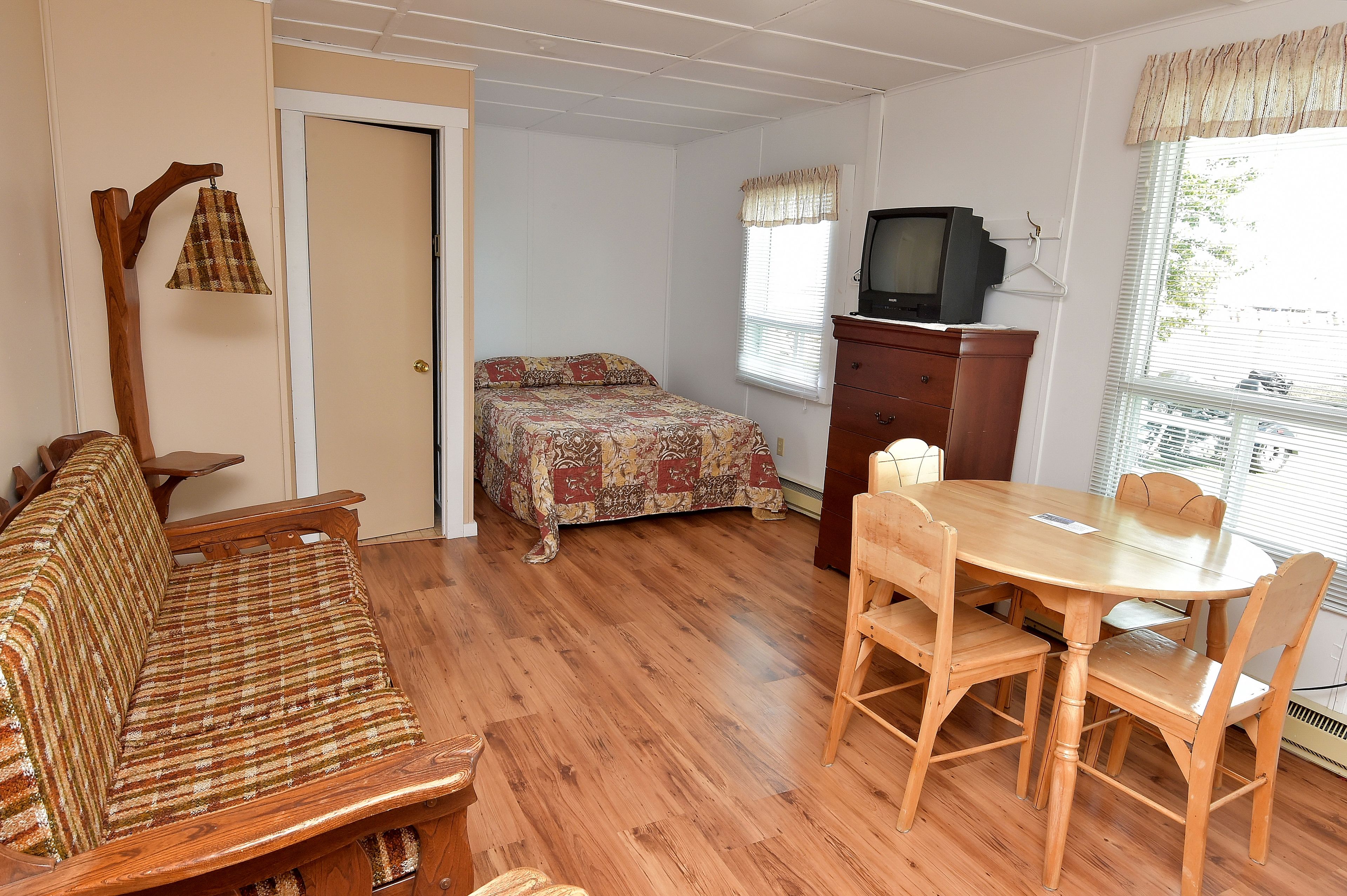 Ein Zimmer mit einer Holzsofa, einem Esstisch mit Stühlen, einem Bett mit einem gemusterten Bettbezug, einem Fernseher auf einer Holzkommode und einem Fenster mit Jalousien.