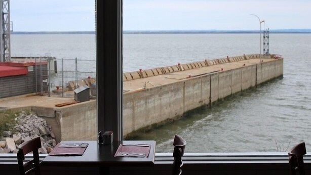 Blick aus einem Fenster auf einen Betonsteg, der sich ins ruhige Wasser erstreckt, mit einer Metallkonstruktion und einem Gebäude mit rotem Dach im Hintergrund.