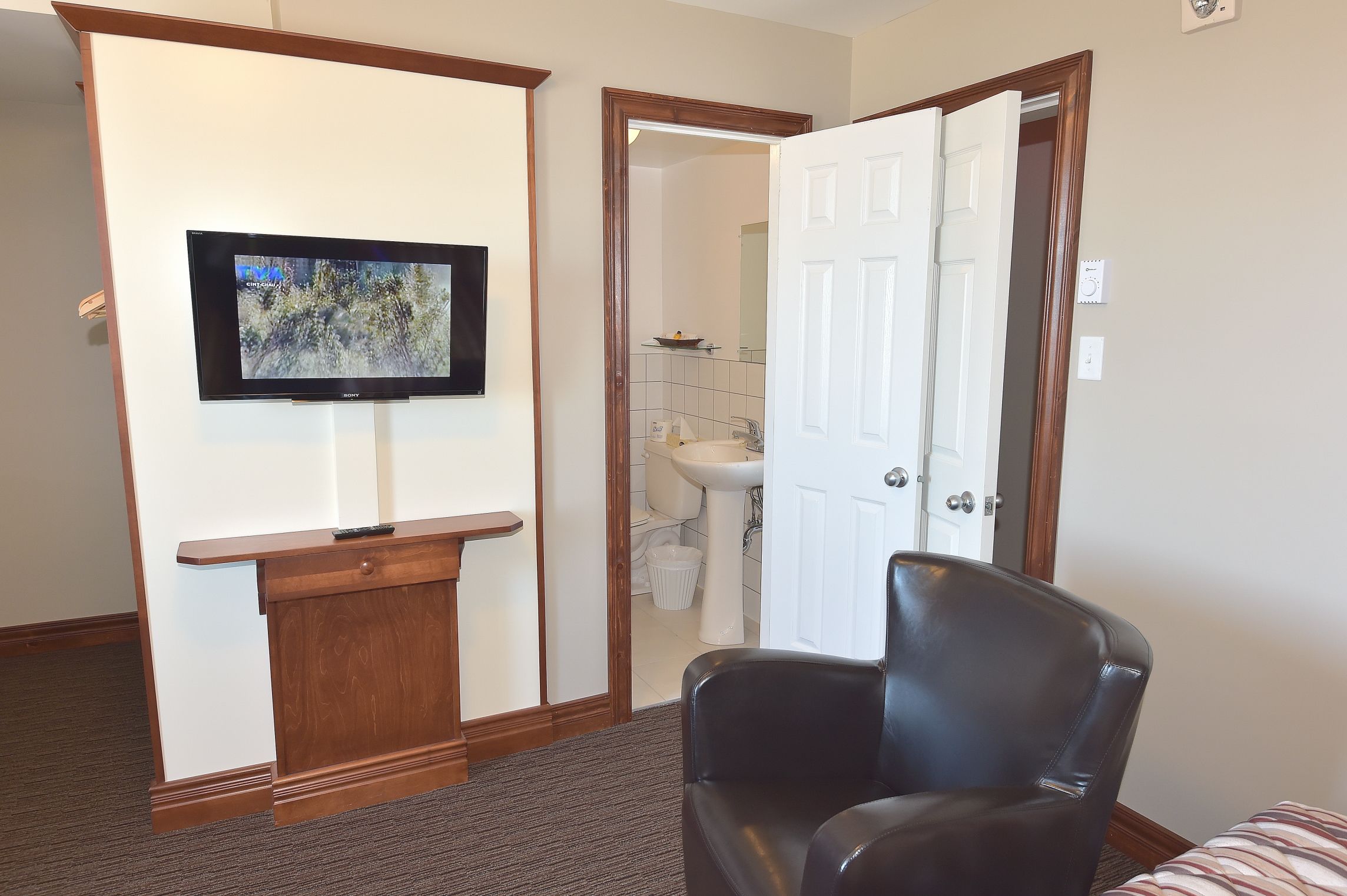 Ein Hotelzimmer mit einem Holztisch, einem an der Wand befestigten Fernseher, einem schwarzen Ledersessel und einem durch eine offene Tür einsehbaren Badezimmer.