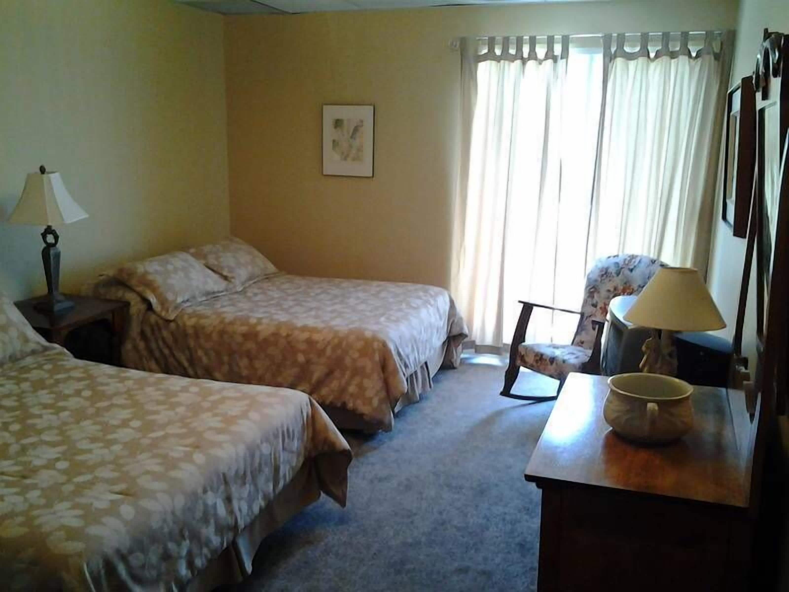 Habitación de hotel con dos camas, una silla, un escritorio con lámpara y una ventana con cortinas.