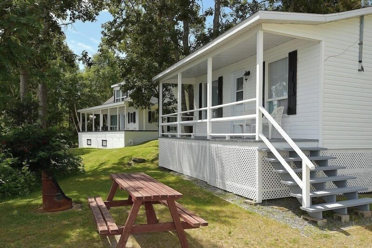 A white cabin with a porch and a picnic table in a grassy area.