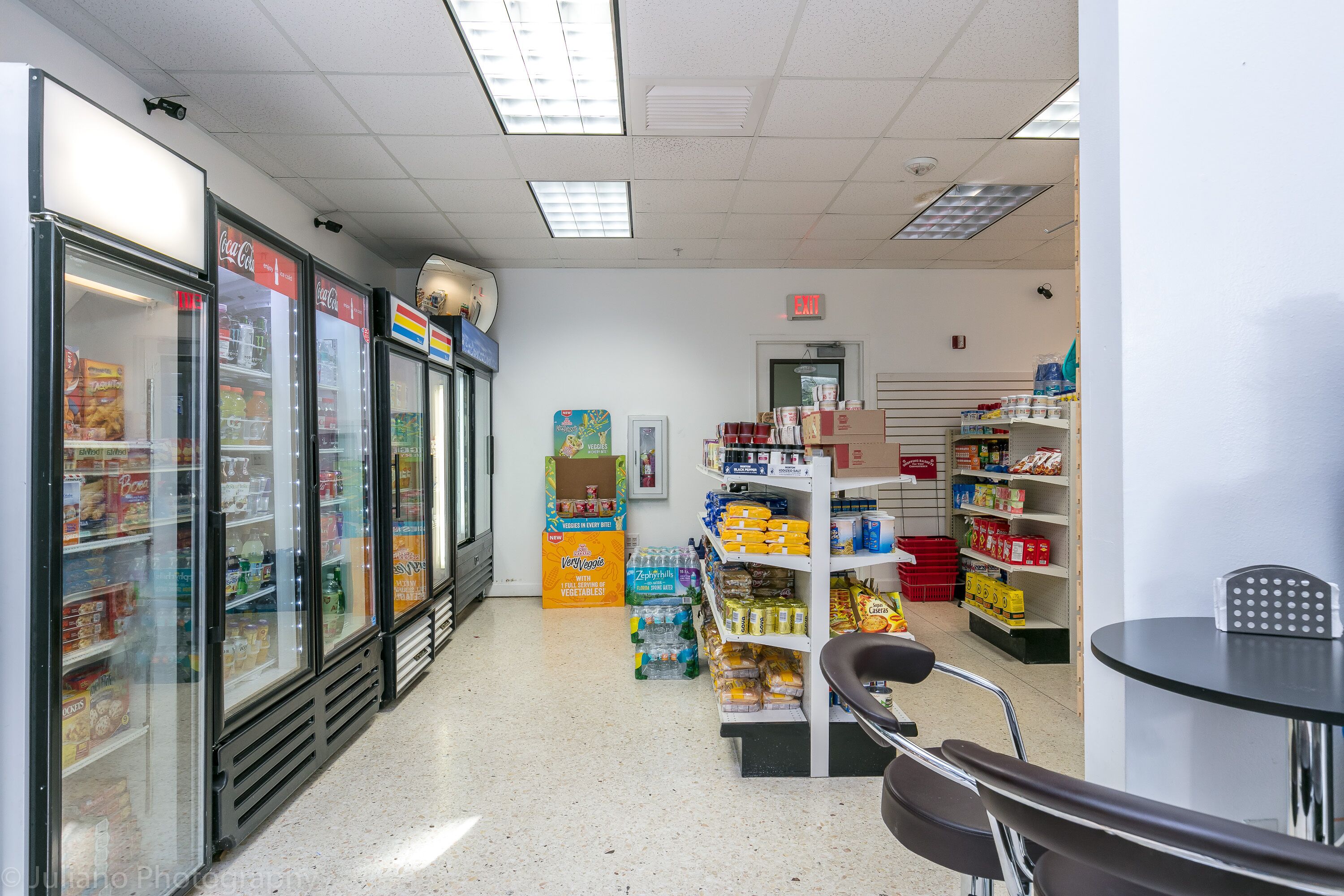 Interior de un supermercado con secciones refrigeradas, estantes llenos de productos varios y un área con mesas.