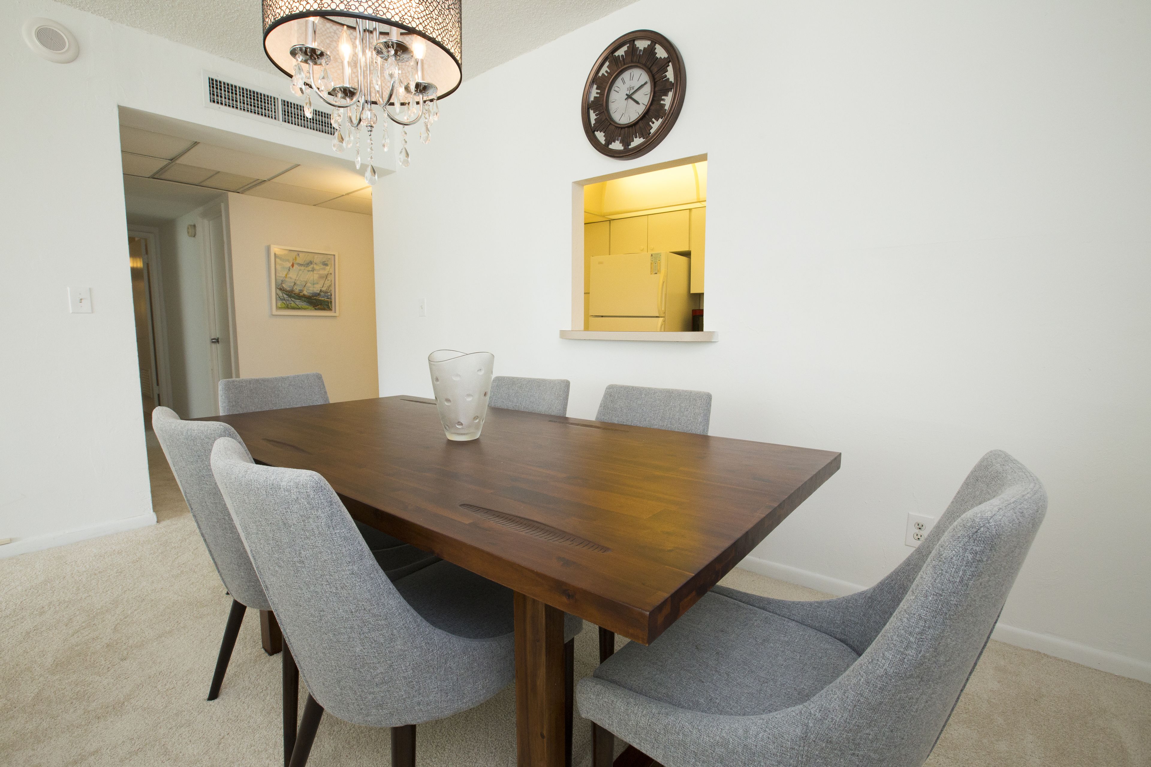 A dining room with a wooden table, grey upholstered chairs, a chandelier, a wall clock, and a mirror.