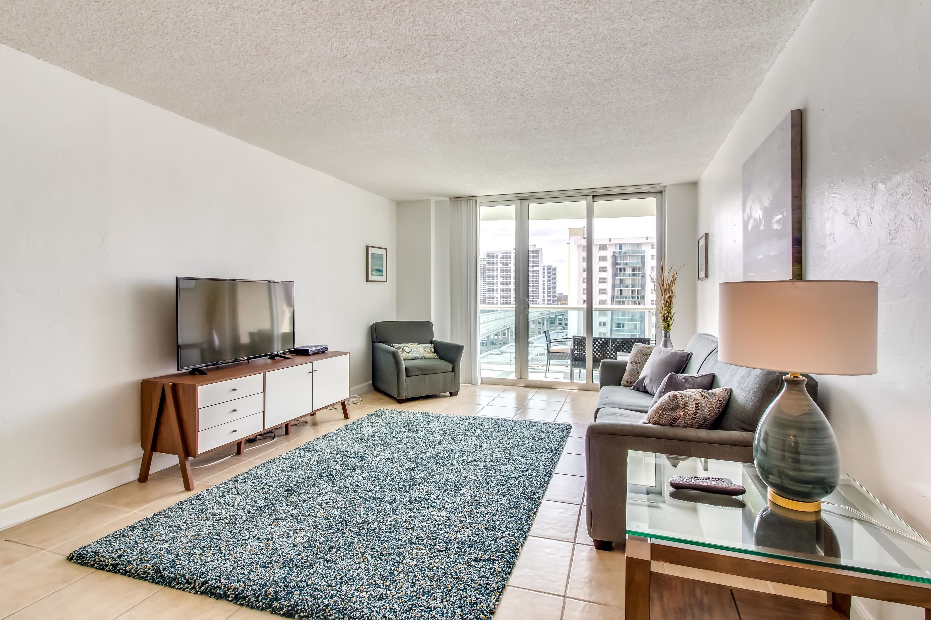 A modern living room with a flat-screen TV, a sofa, a glass coffee table, and a large window offering a city view.