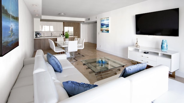 A modern living room with a glass coffee table, white sofas, and a flat-screen TV mounted on the wall. - Ocean Reserve Luxury Condos Across from Sunny Isles Beach (Sunny Isles Beach)