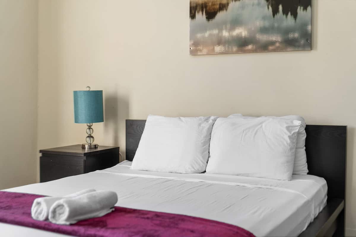 Een net opgemaakt bed met witte lakens en een paarse deken, een blauwe bedlamp en een schilderij van een natuurscène aan de muur.