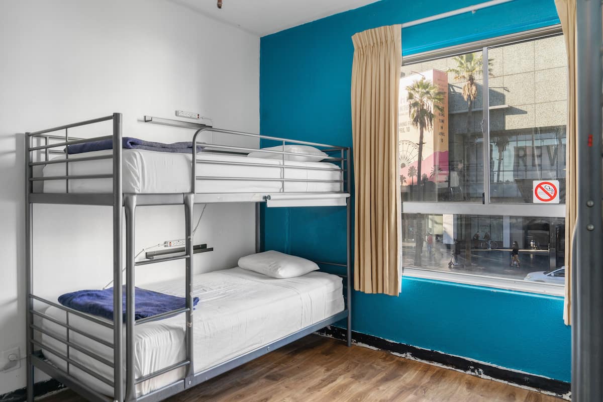 Een slaapkamer met een stapelbed, een raam met gordijnen, een houten vloer en uitzicht op de straat.