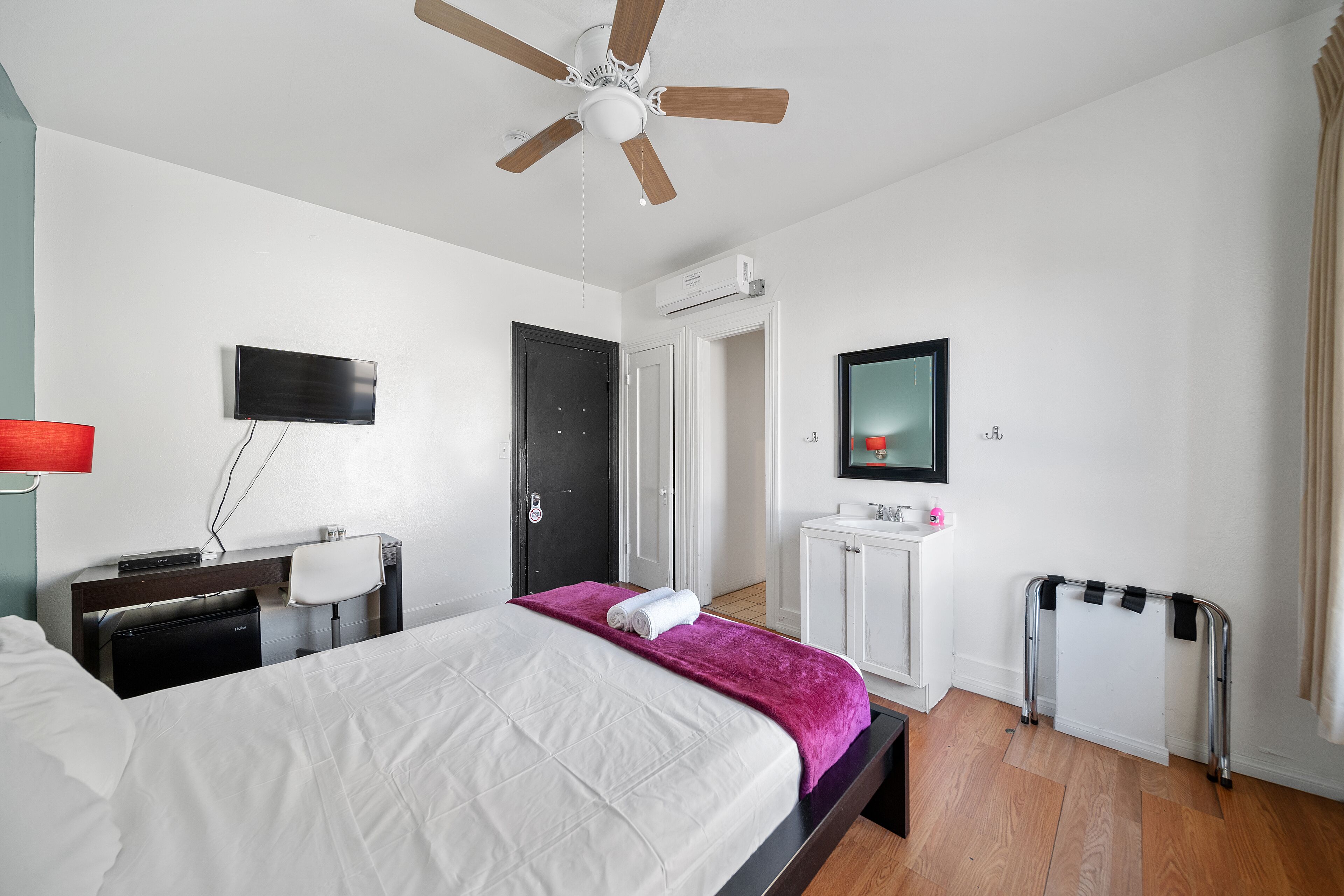 Ein Hotelzimmer mit Bett, Schreibtisch mit Stuhl, Fernseher, Badezimmer und Deckenventilator.