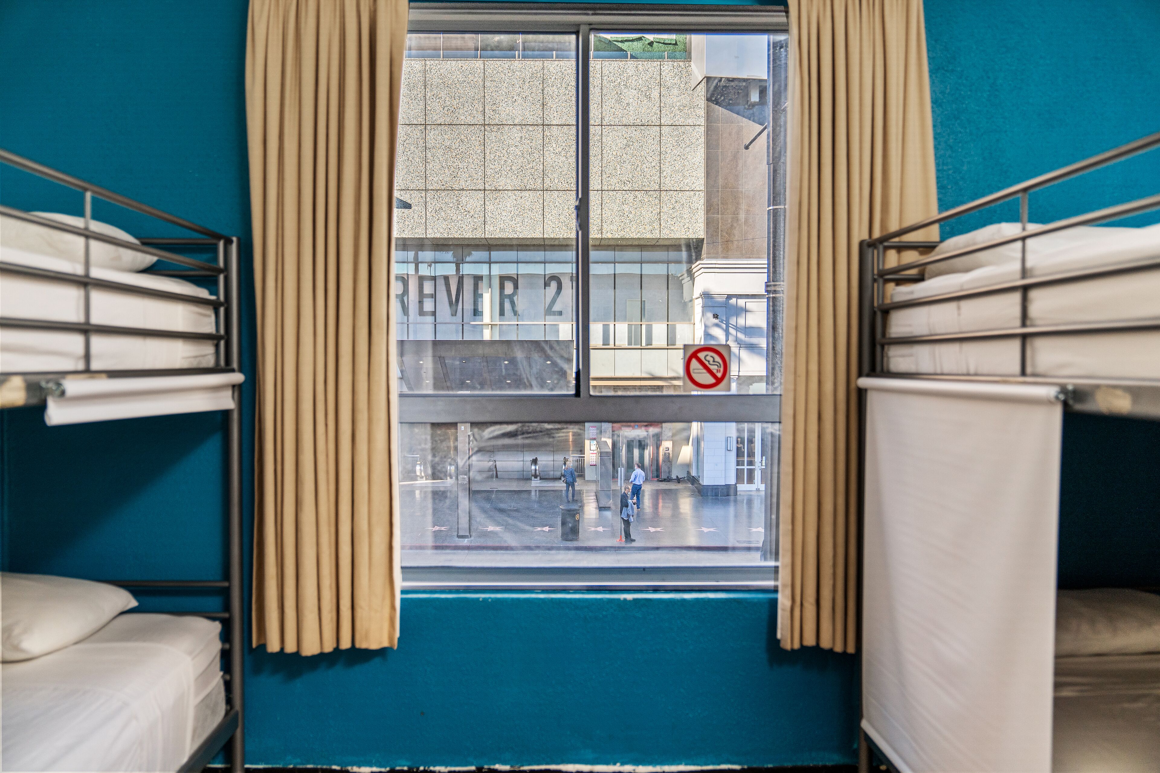 A room with bunk beds, a window with curtains, and a view of the street outside.