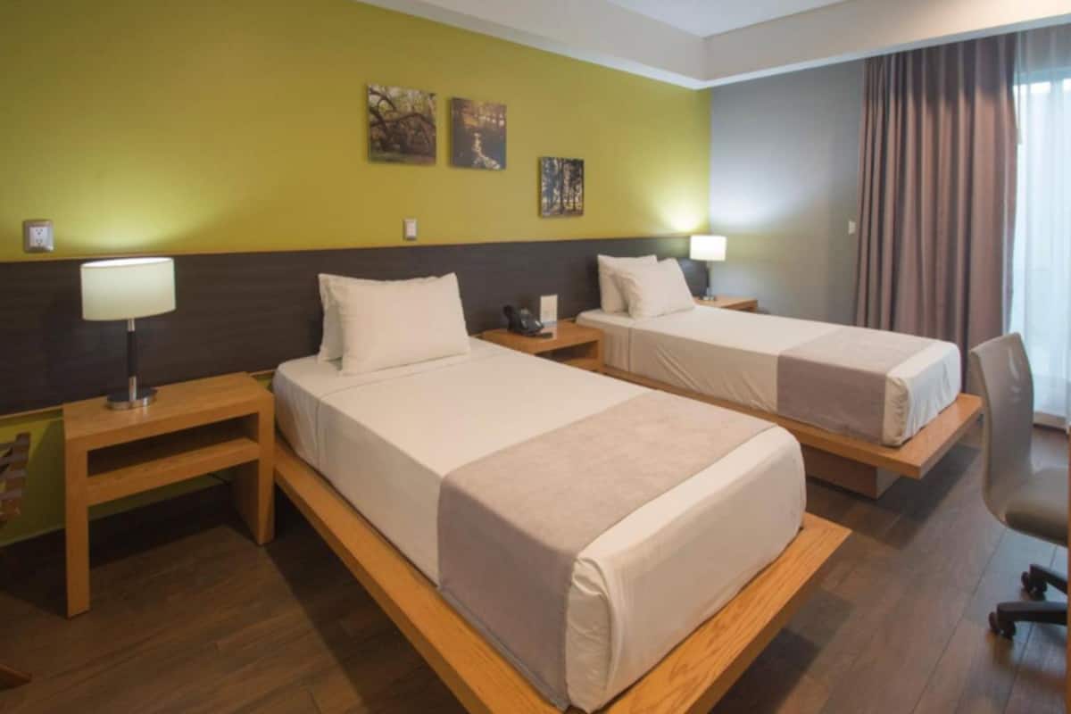 A hotel room with two beds, wooden headboards, a nightstand with a lamp, and artwork on the wall.