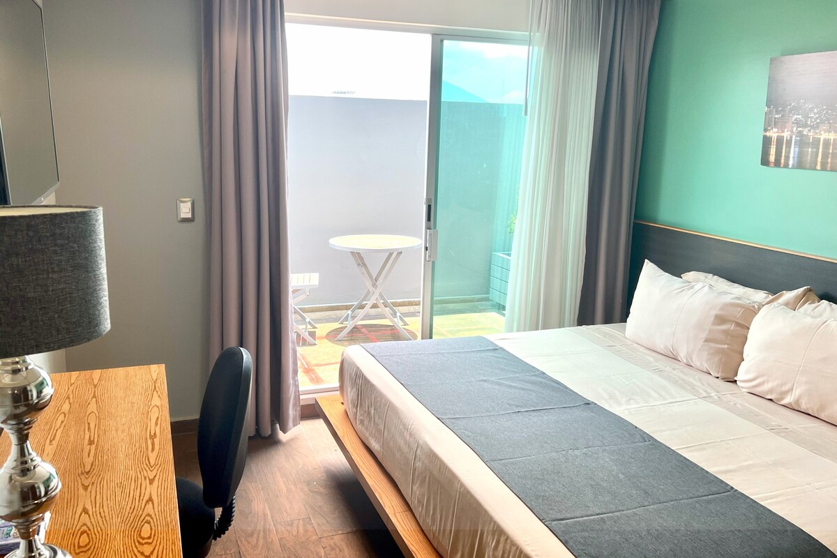Habitación de hotel con cama, escritorio, silla, lámpara y vistas al exterior.