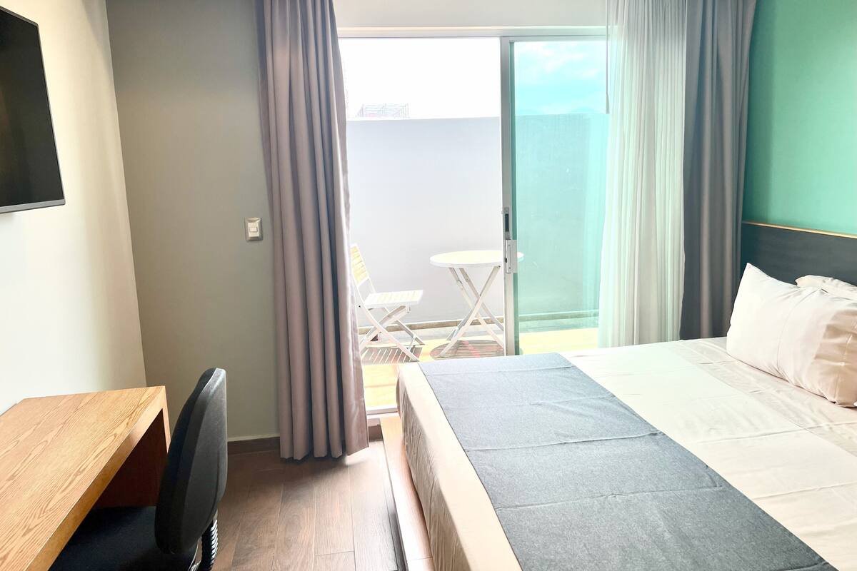 A hotel room with a bed, a desk, a chair, a small table, and a view of the outside.