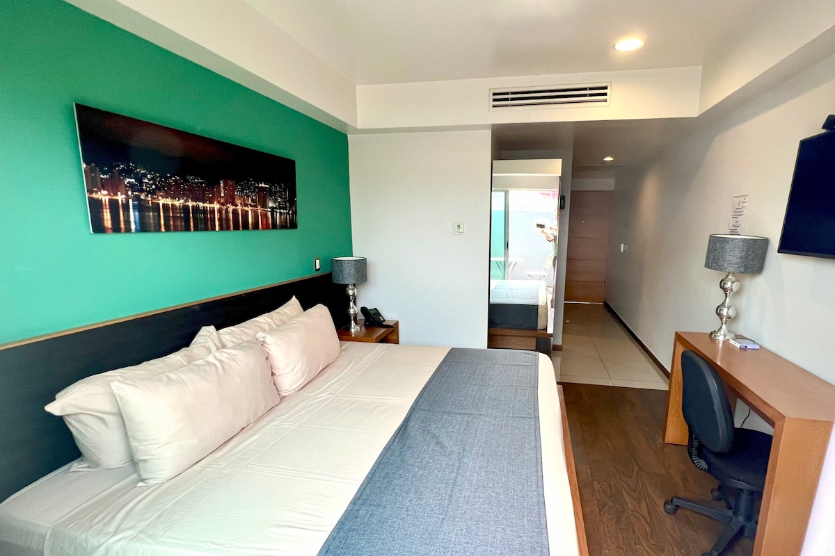 A hotel room with a large bed, a desk, a chair, a desk lamp, a bedside table, and a painting on the wall.