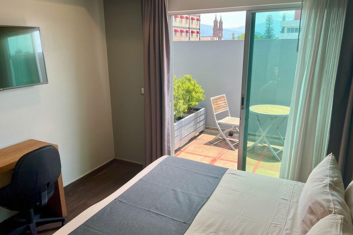 A hotel room with a bed, a desk, a chair, a small table, a balcony with a chair and a table, and a view of buildings and trees.