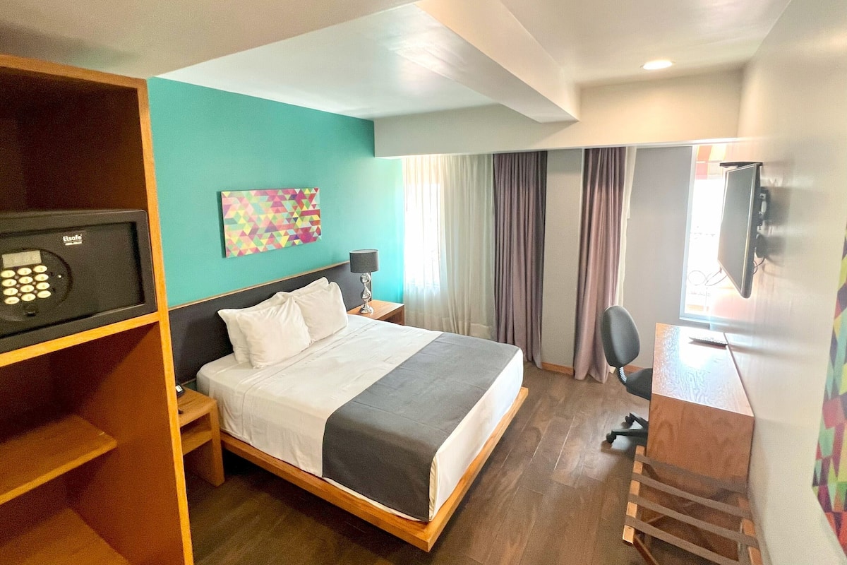 A hotel room with a bed, a desk, a TV, a safe, and a colorful artwork on the wall.