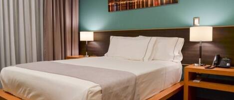 Habitación de hotel con cama, dos mesitas de noche, una lámpara y un cuadro de un paisaje urbano en la pared.