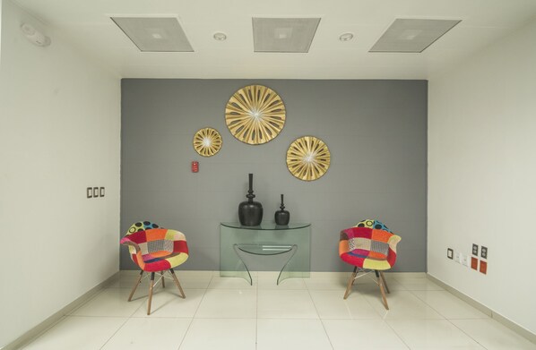 Quarto moderno com parede em cinza, três peças decorativas na parede, mesa de console de vidro, duas cadeiras coloridas e teto com iluminação embutida.