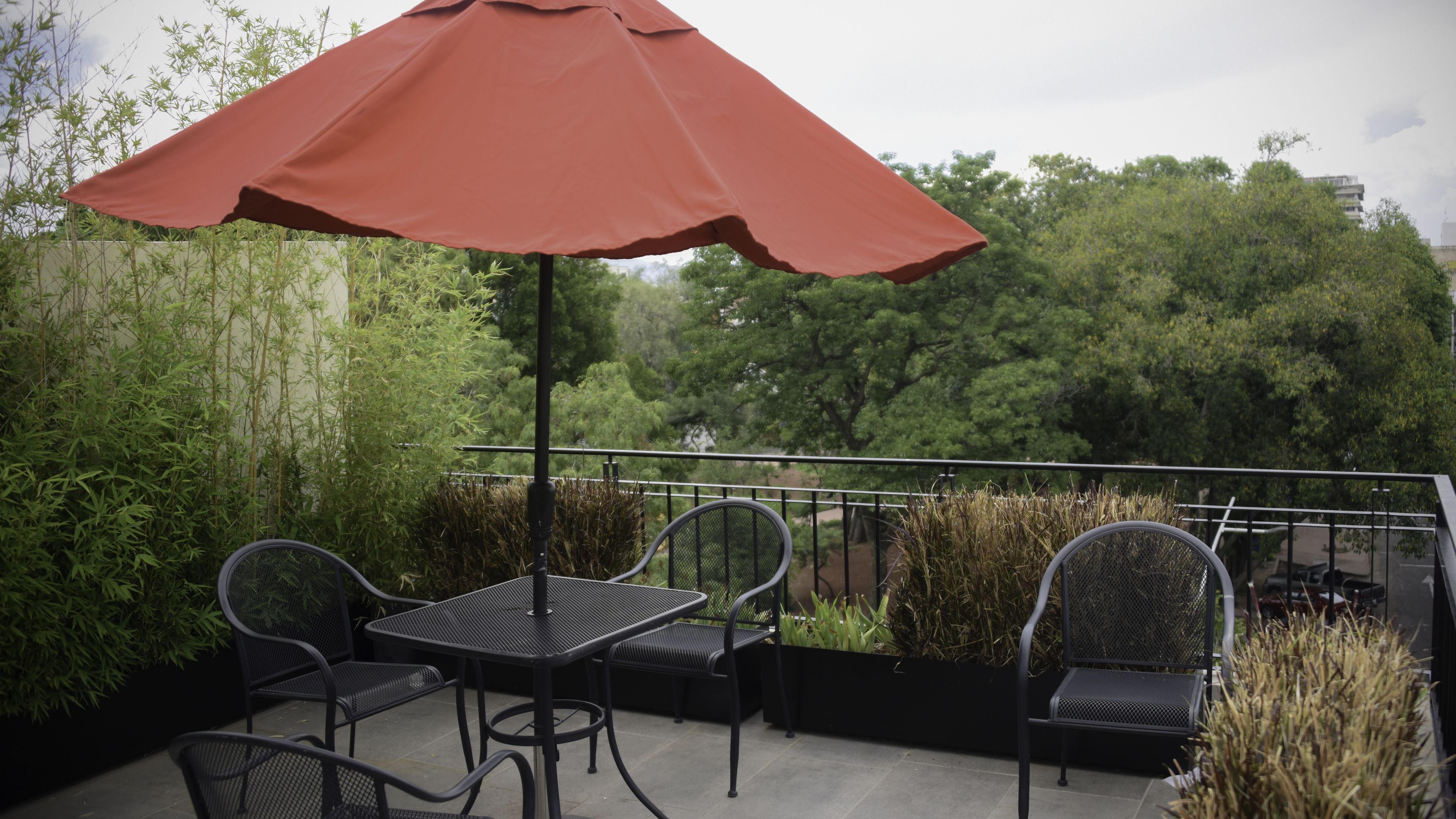 Une terrasse extérieure avec une table et des chaises, un parasol rouge et une vue sur les arbres et les bâtiments.