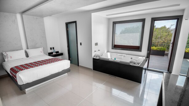 Un dormitorio moderno con una cama grande, una bañera integrada y un balcón con vistas a árboles.