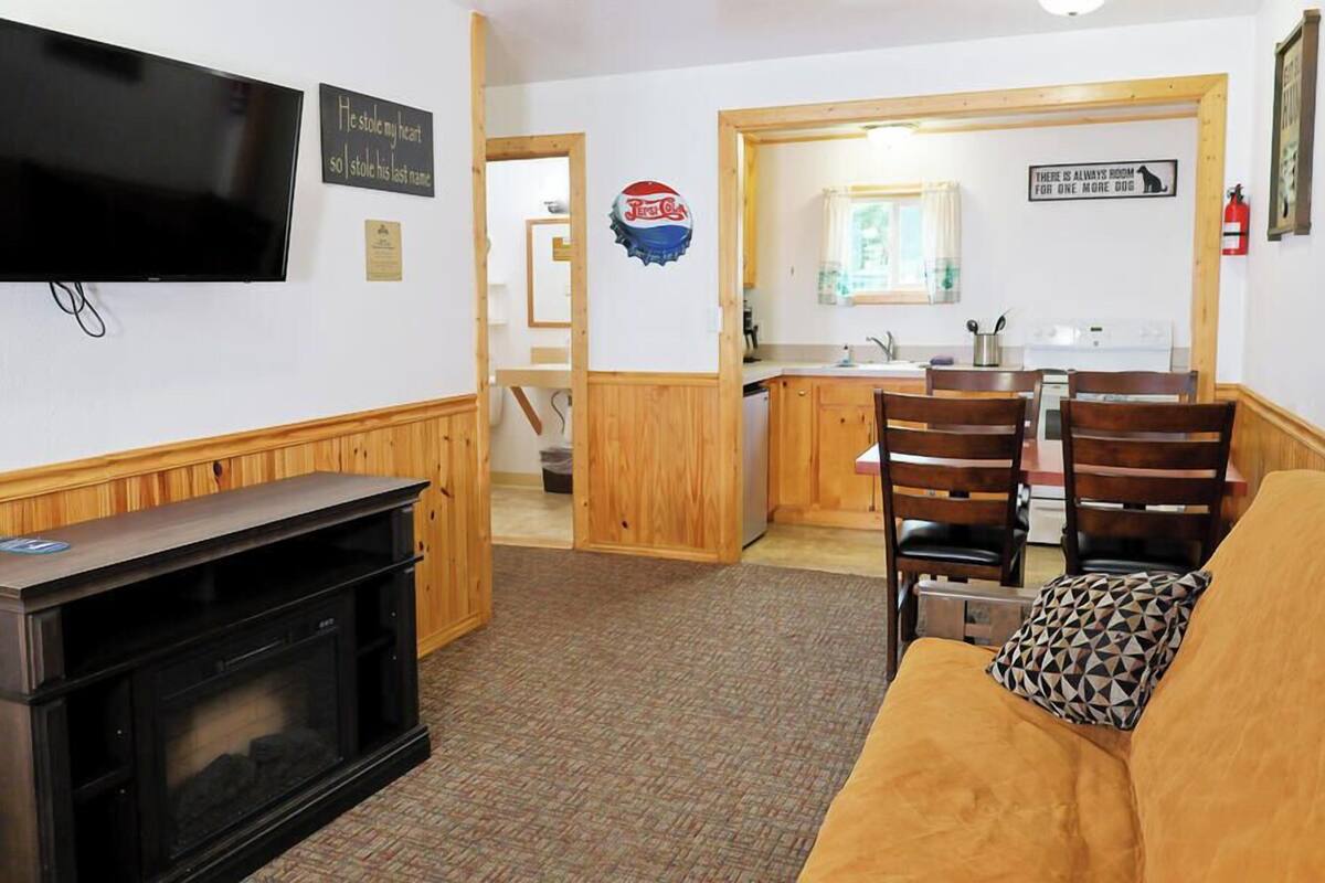 A living room with a TV, wooden furniture, a kitchen in the background, and a sign on the wall.