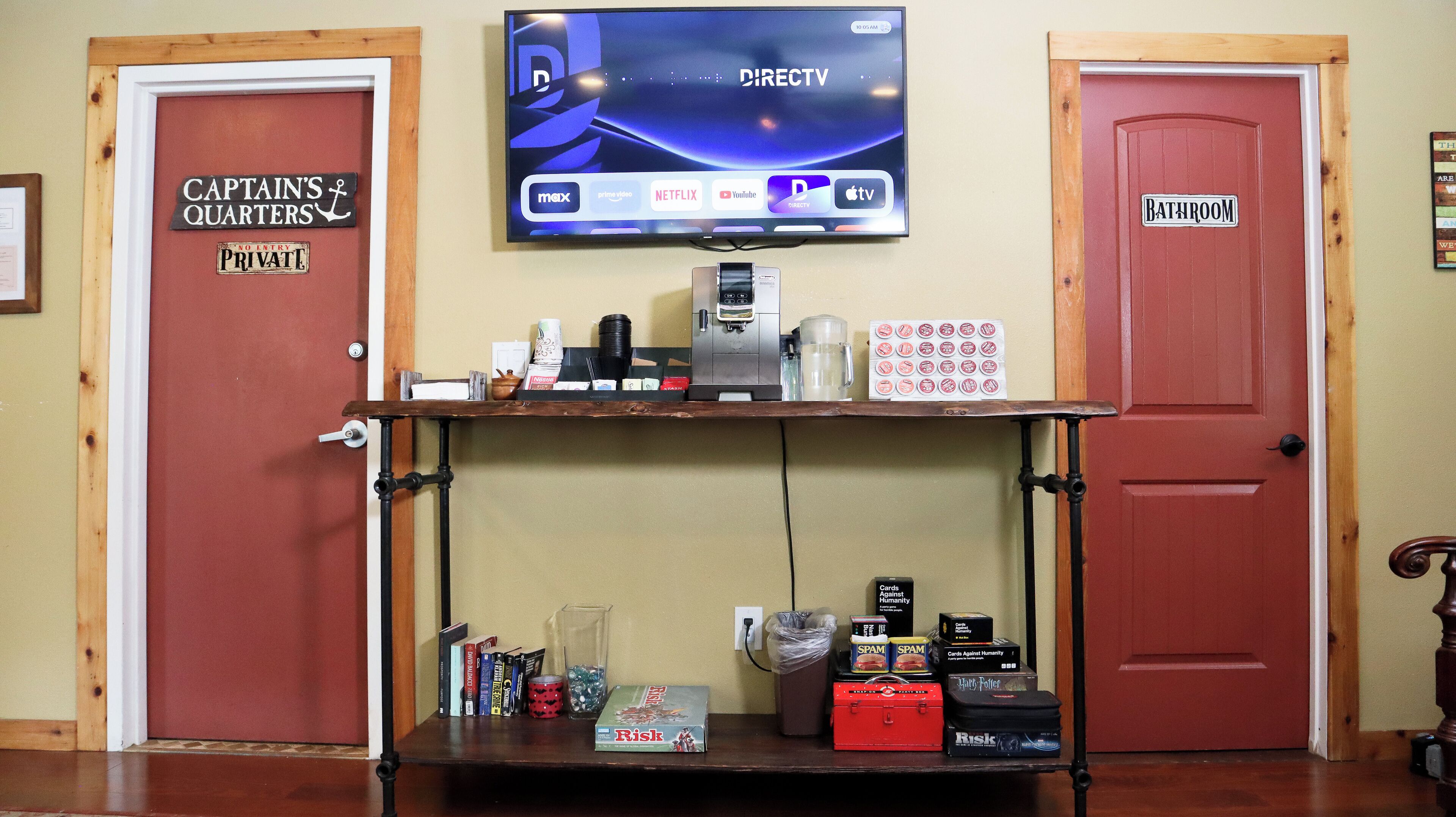 A room with a television displaying DIRECTV channels, a coffee machine, and a shelf with books and games.