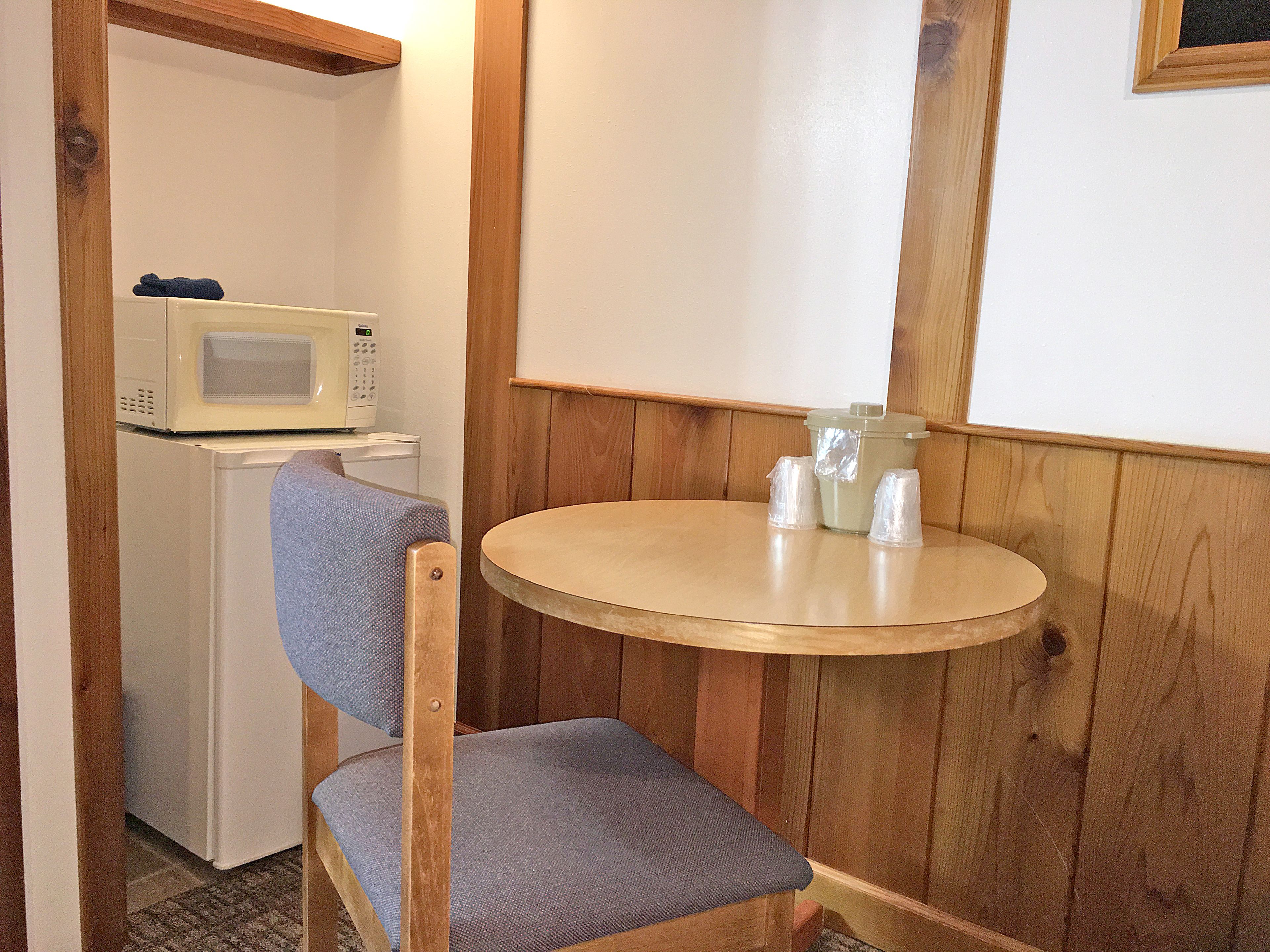 A small kitchenette with a microwave, refrigerator, and a wooden chair with a blue cushion.