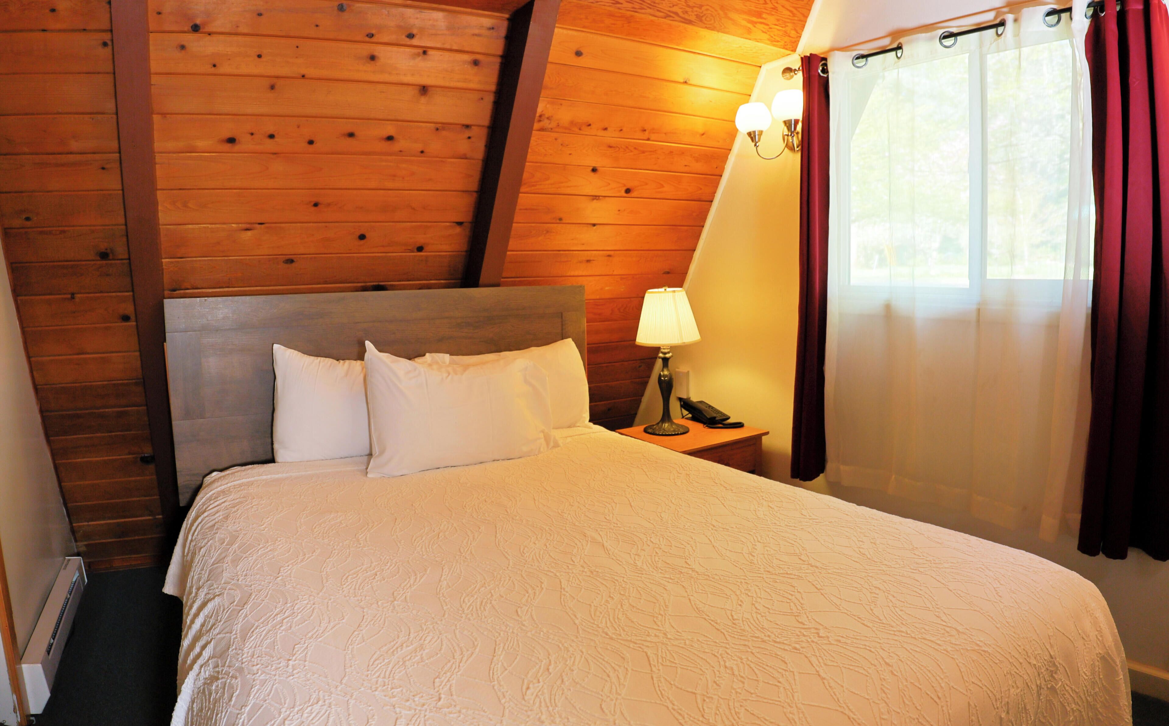 Un dormitorio con techo de madera, una cama con ropa de cama blanca, una mesita de noche con lámpara y una ventana con cortinas.