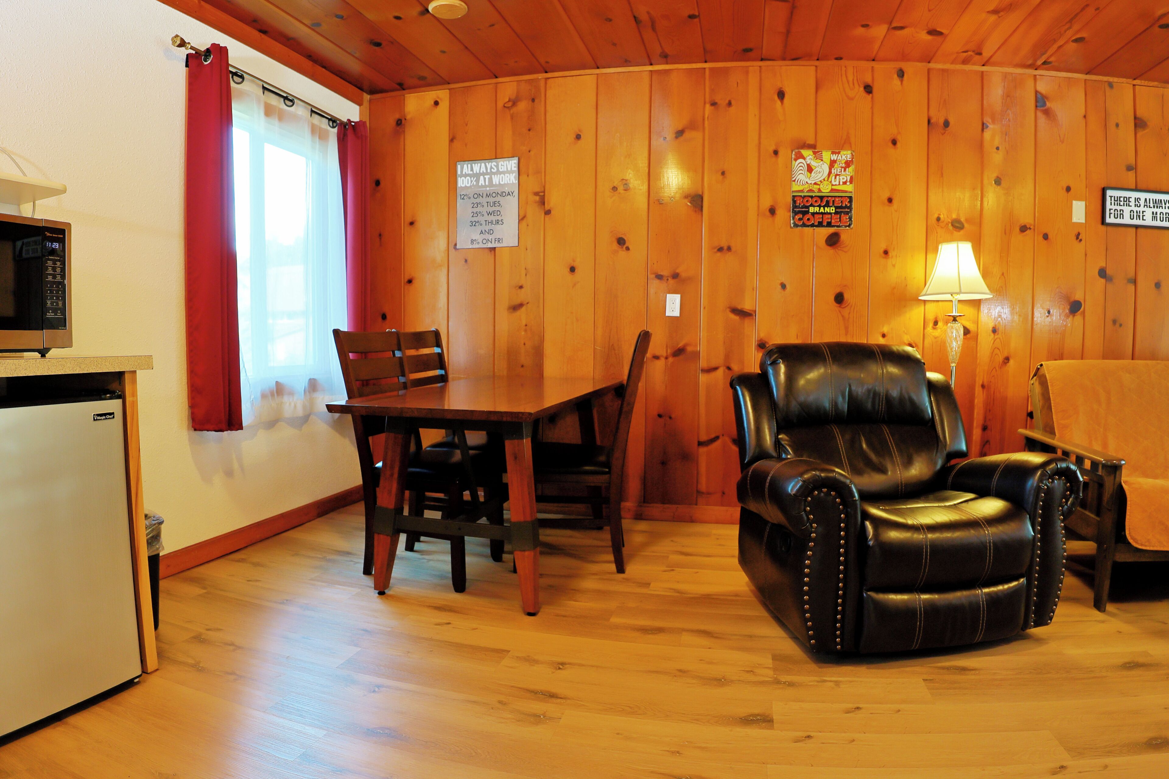 Ein Zimmer mit Holzwänden, einem Esstisch, Stühlen, einem Ledersessel, einer Lampe und einem Kühlschrank.