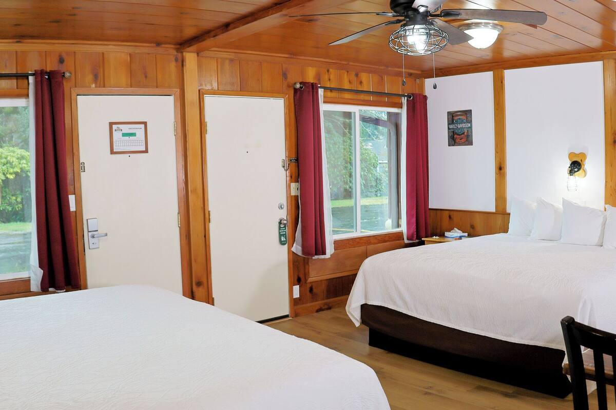 A King Quad room with two beds, wooden walls, a ceiling fan, and large windows with curtains.