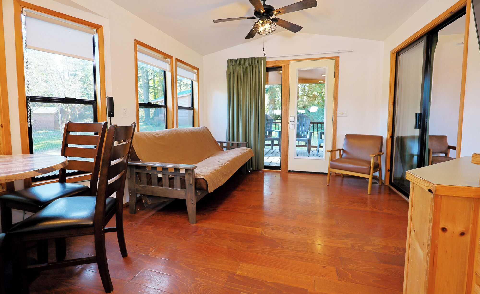 A room with a wooden floor, a sofa, a dining table with chairs, a ceiling fan, and large windows.