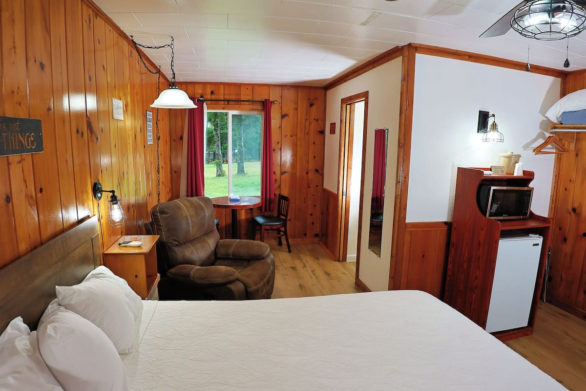 A wooden room with a bed, a chair, a small table, a microwave, and a refrigerator.