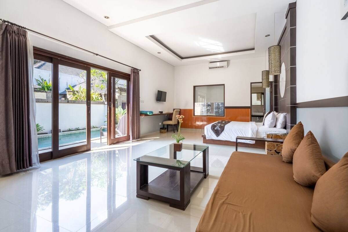 A modern hotel room with a large bed, a glass coffee table, a sofa, a TV, and a view of a swimming pool.