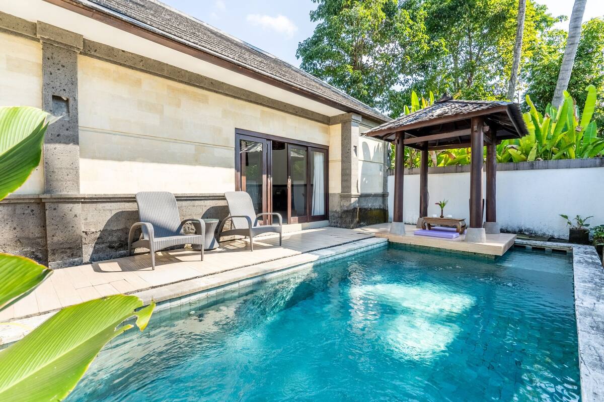 A swimming pool with lounge chairs and a covered seating area, surrounded by a stone wall and tropical plants.