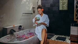 Una persona vestida con bata sentada junto a una bañera grande llena de agua y pétalos de rosa, con un cuenco de pétalos de rosa y varios productos de baño en el suelo.