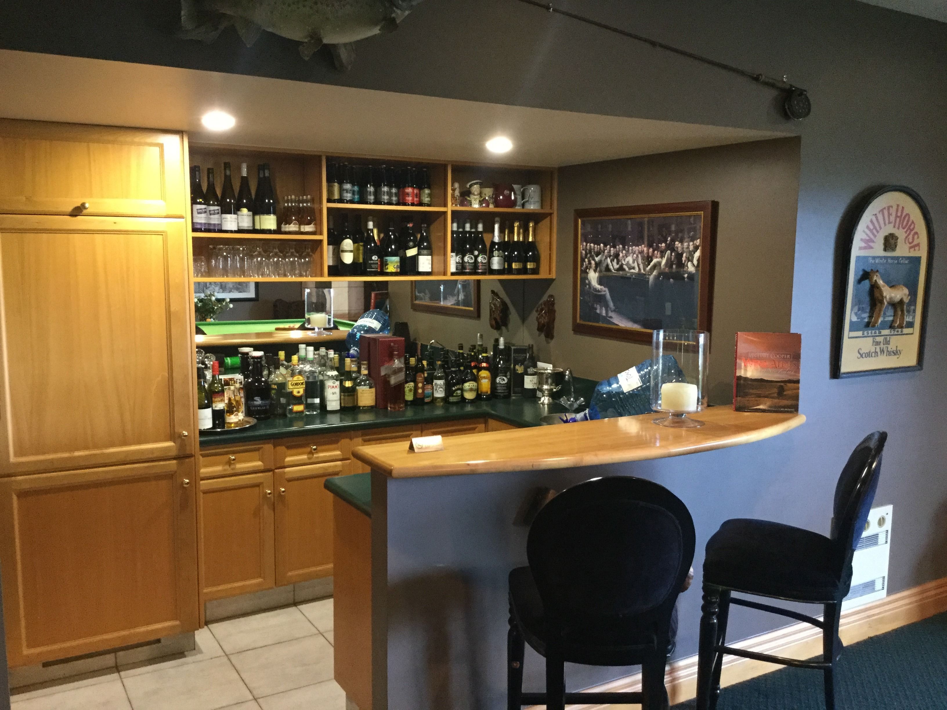 A bar area with shelves of liquor bottles, a bar counter, two chairs, and a framed picture on the wall.