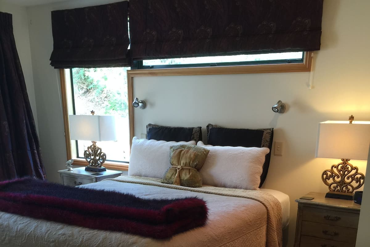 Een slaapkamer met een groot bed, een nachtkastje met een lamp, een stoel en een raam met gordijnen.