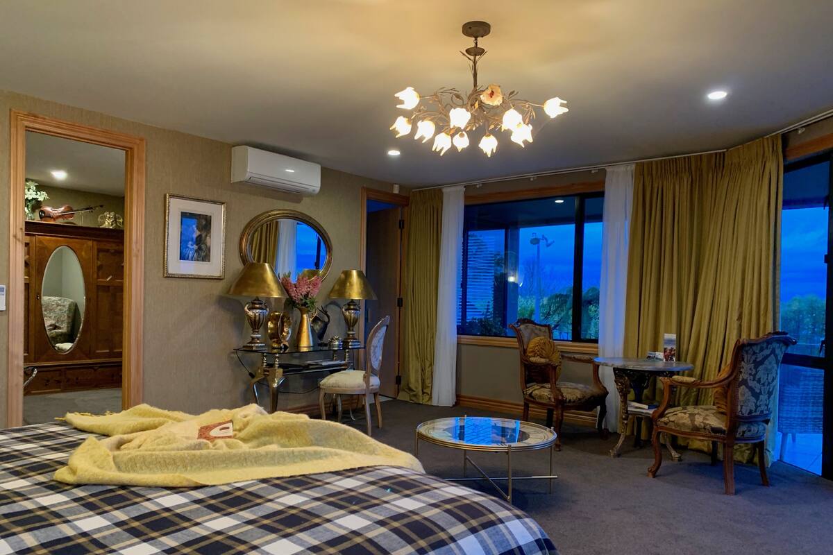 Hotelkamer met een groot bed, een bureau, twee stoelen, een ronde tafel, een spiegel en een raam met gordijnen.