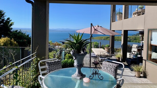 Una mesa de vidrio redonda con un florero decorativo y una única piedra rosa, rodeada de sillas estampadas, con vistas panorámicas del mar y las montañas.