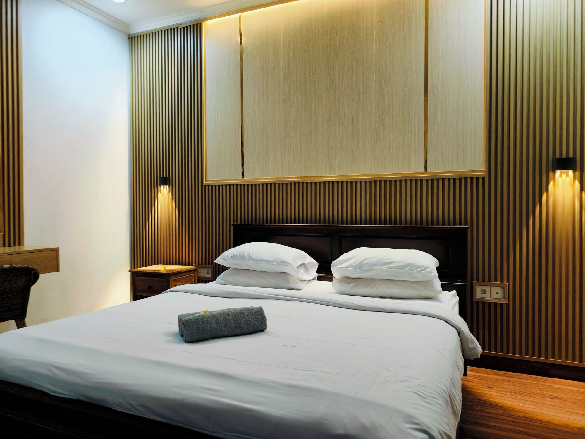 A hotel room with a large bed, two bedside tables, a wooden headboard, and wall-mounted lighting.