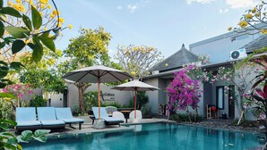 A swimming pool with lounge chairs and umbrellas, surrounded by lush greenery and flowering plants. - Ashana Hotel Uluwatu (Pecatu)