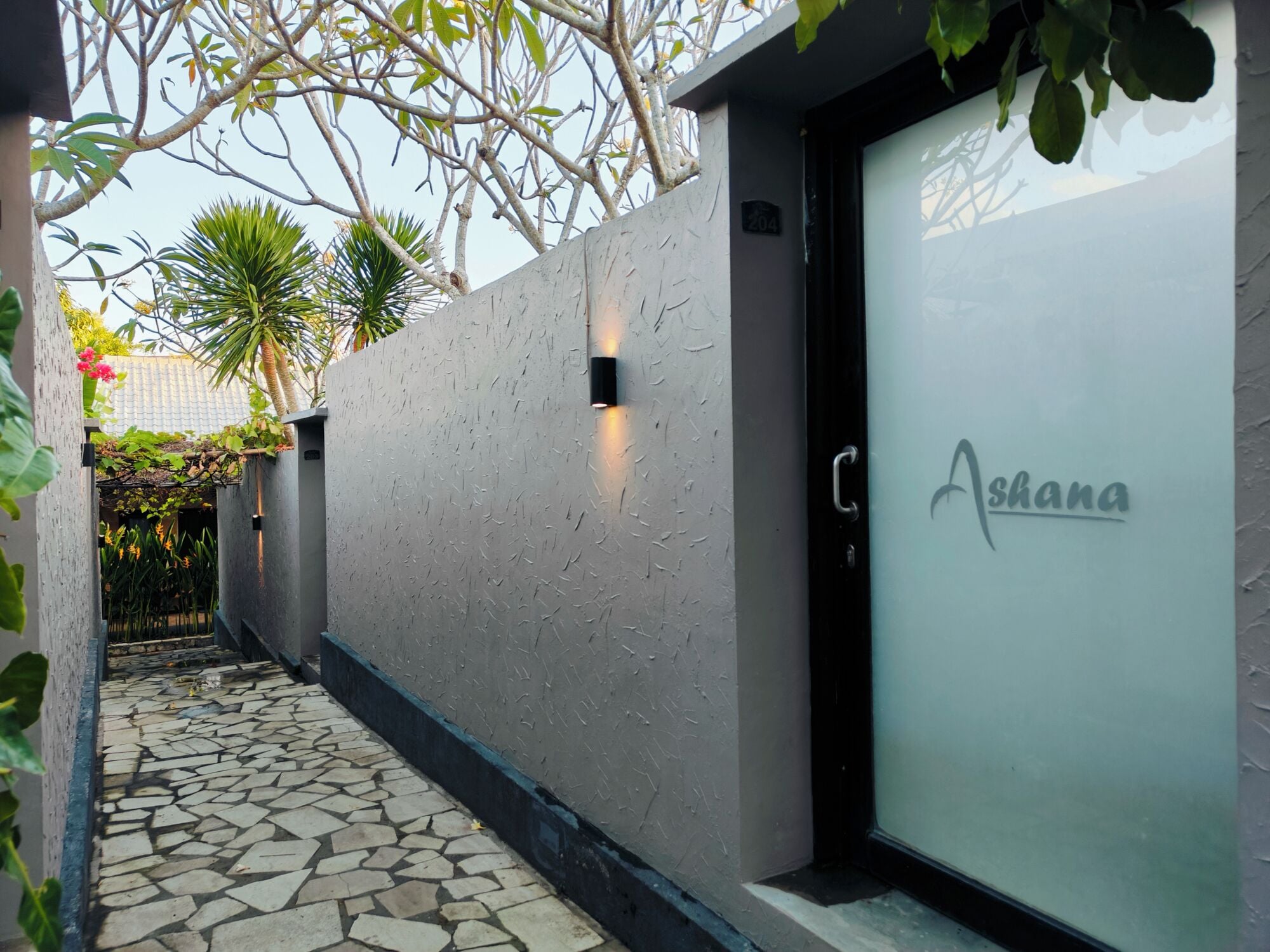 A modern building facade with a textured wall, a door with the name 'Ashana' on it, and a stone pathway leading to the entrance.