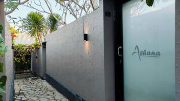 A modern building facade with a textured wall, a door with the name 'Ashana' on it, and a stone pathway leading to the entrance.