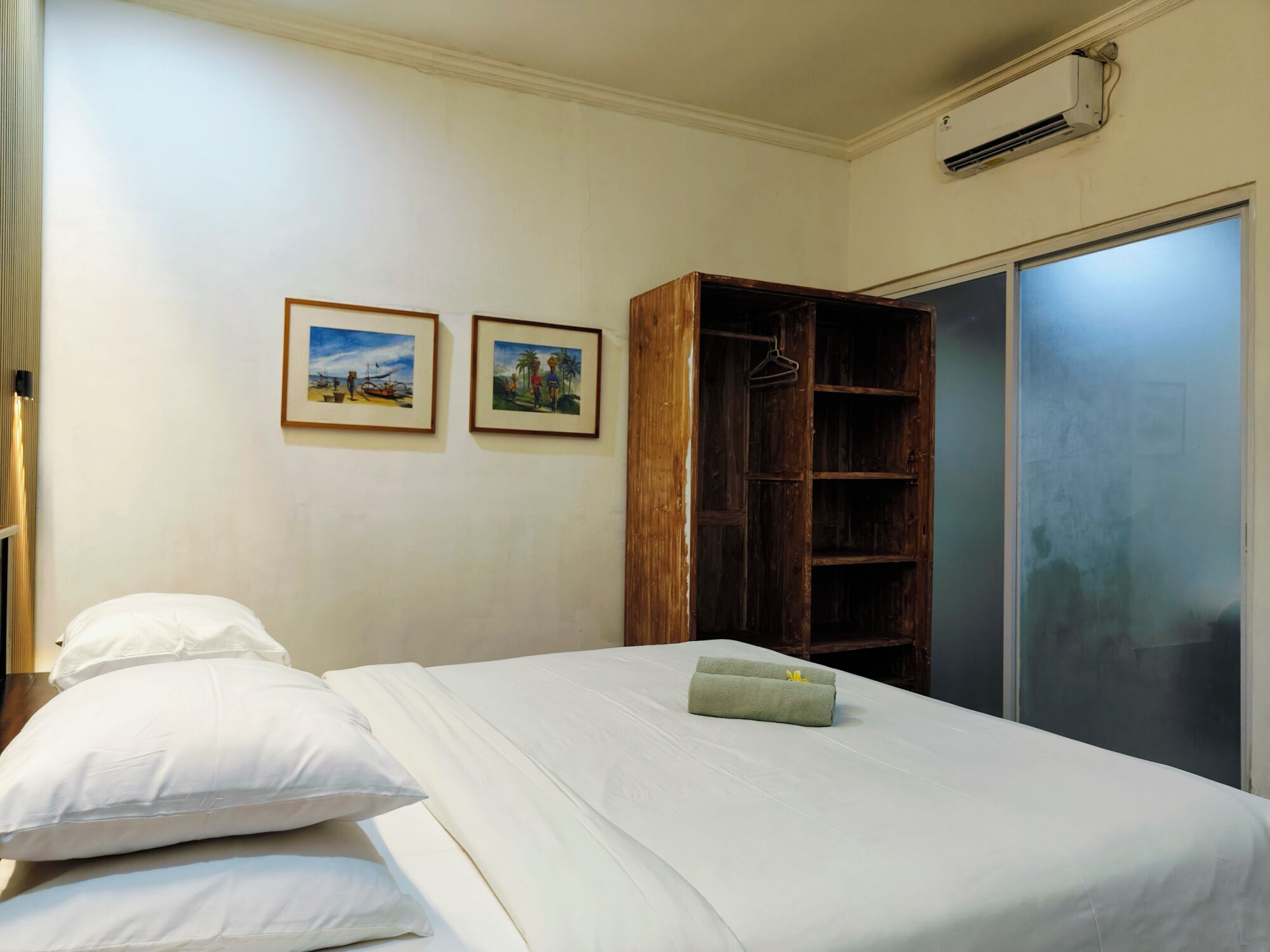 A hotel room with a bed, two pillows, a wooden wardrobe, and two framed pictures on the wall.