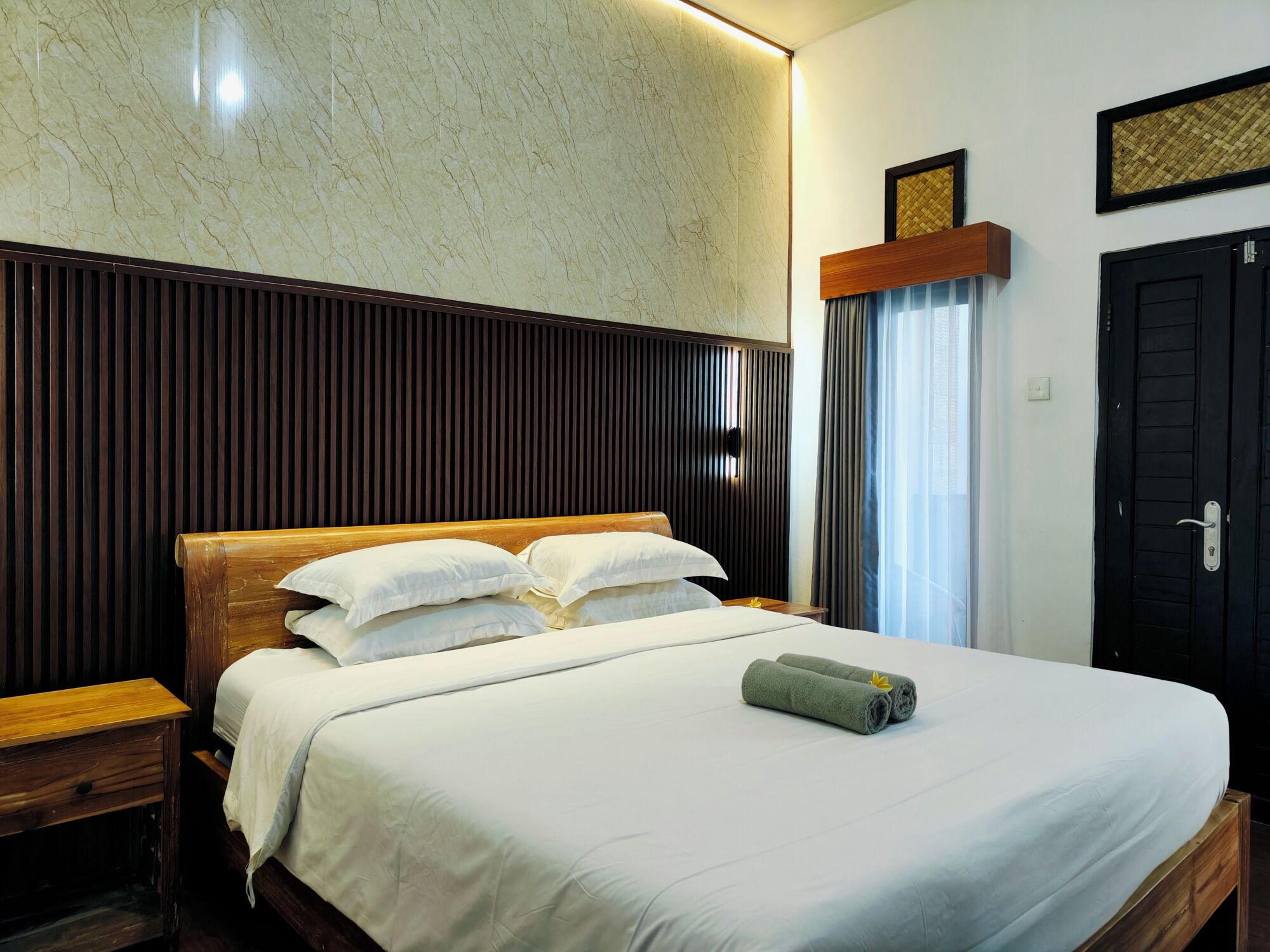 Habitación de hotel con cabecera de madera, cama con sábanas blancas, mesita de noche de madera y estante fijado a la pared.