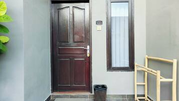 A dark wooden door with a handle, a small window with a curtain, a trash bin, and a wooden ladder leaning against the wall.
